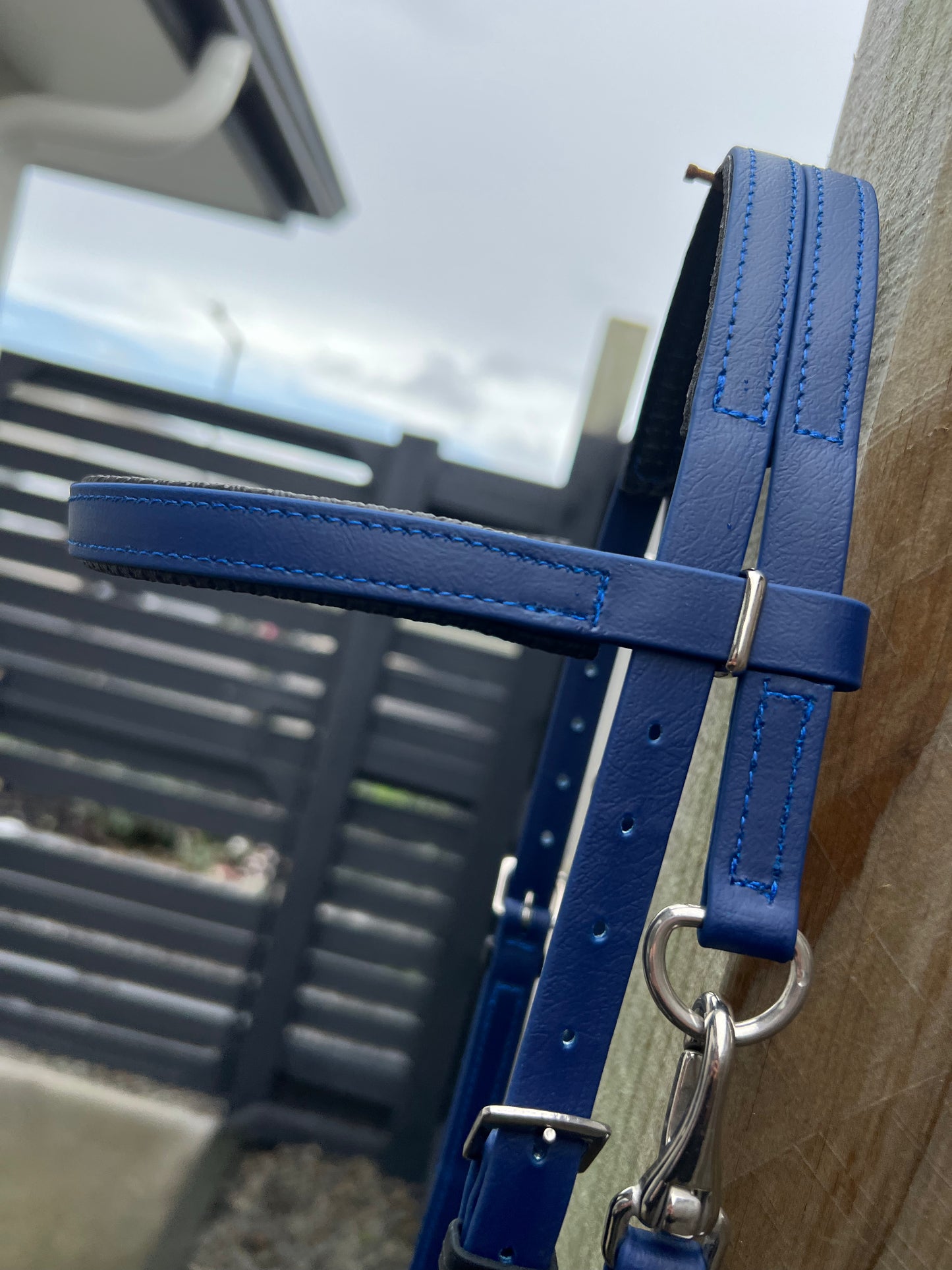 A close-up of the Padded Race Bridle - Navy hanging on a wooden post, reflecting the durability synonymous with LS Equestrian. The background features a gray fence and a cloudy sky.