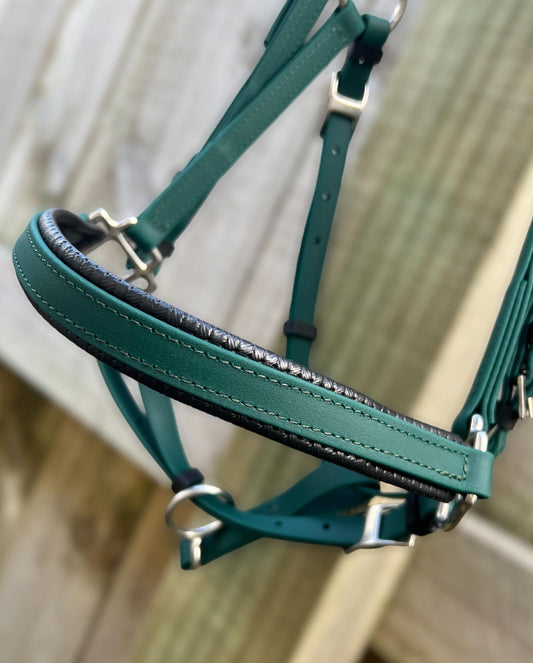 Close-up of the Padded LS Deux Bridle - Hunter Green from LS Equestrian, featuring black padding, hanging in front of a wooden fence. This bridled design features silver metal rings and adjustable straps, making it ideal for endurance riding.