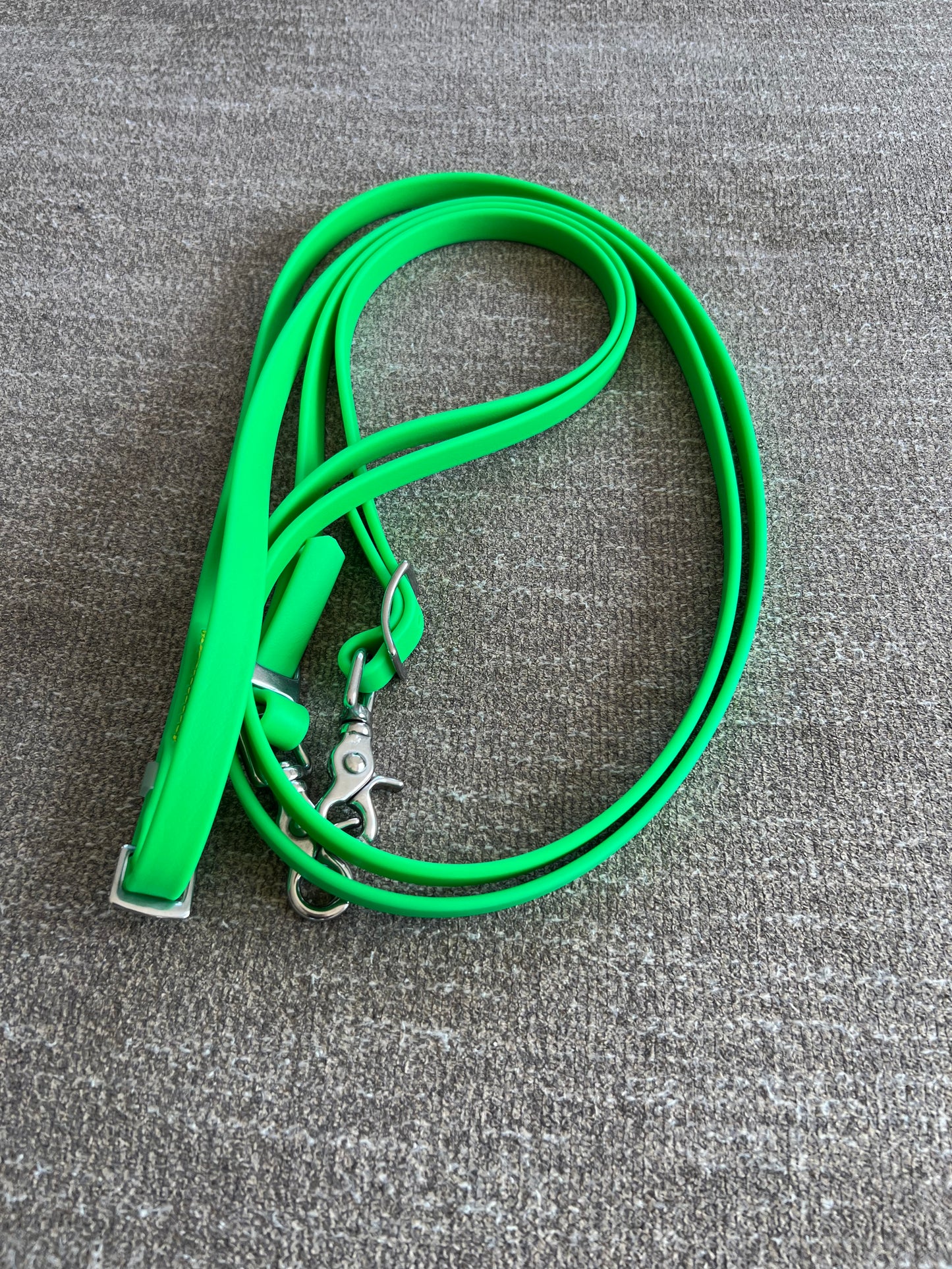 A neon green Reins - Full BioThane from LS Equestrian, featuring stainless steel fittings, is coiled onto a gray textured surface, highlighting its durable construction.