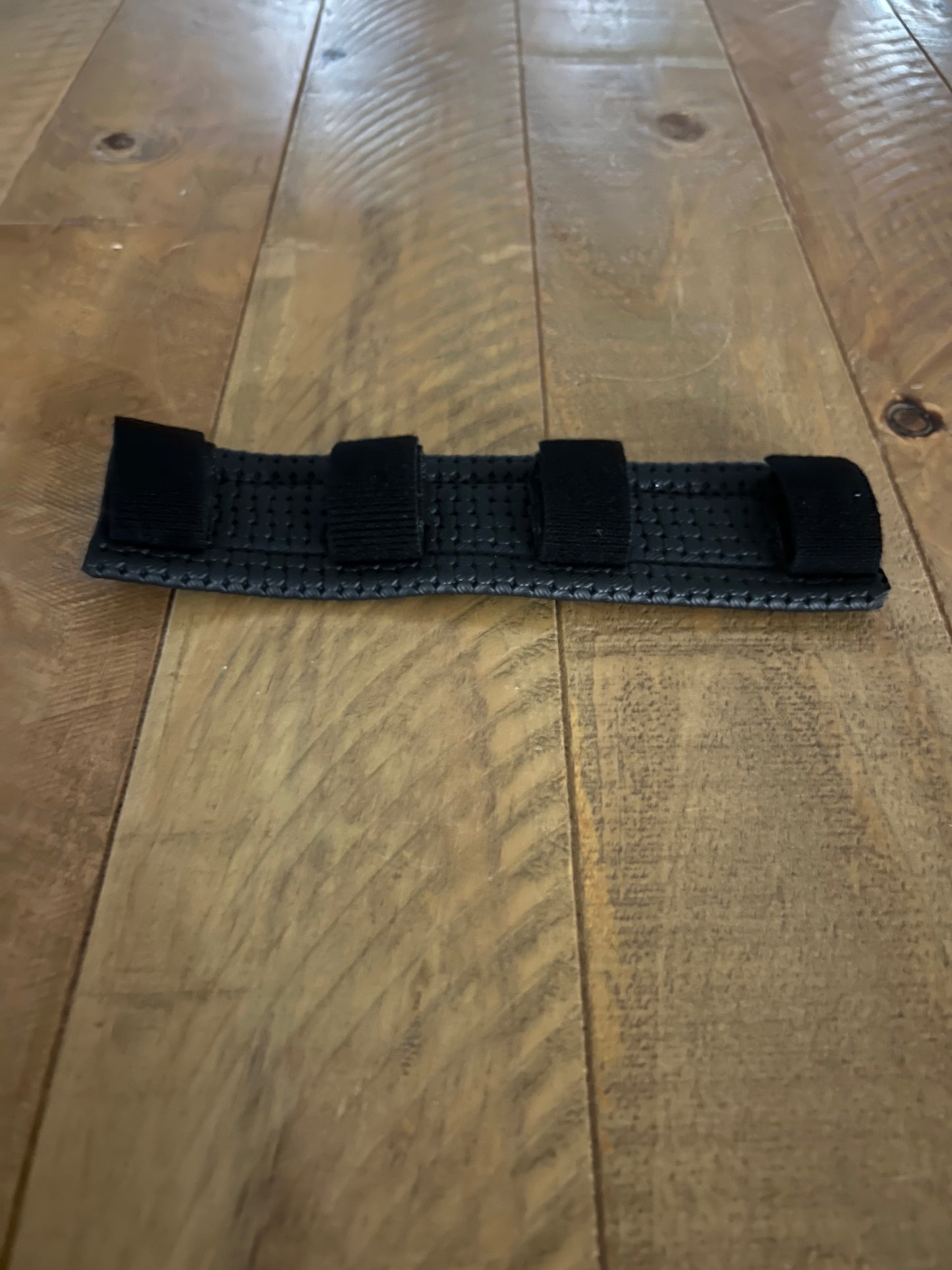 The Poll Padding - Design your own by LS Equestrian, featuring a black fabric strap with Sympa Nova padding and four loops, is displayed on a wooden surface. The textured strap is neatly arranged, with lighting accentuating the wood's grain.