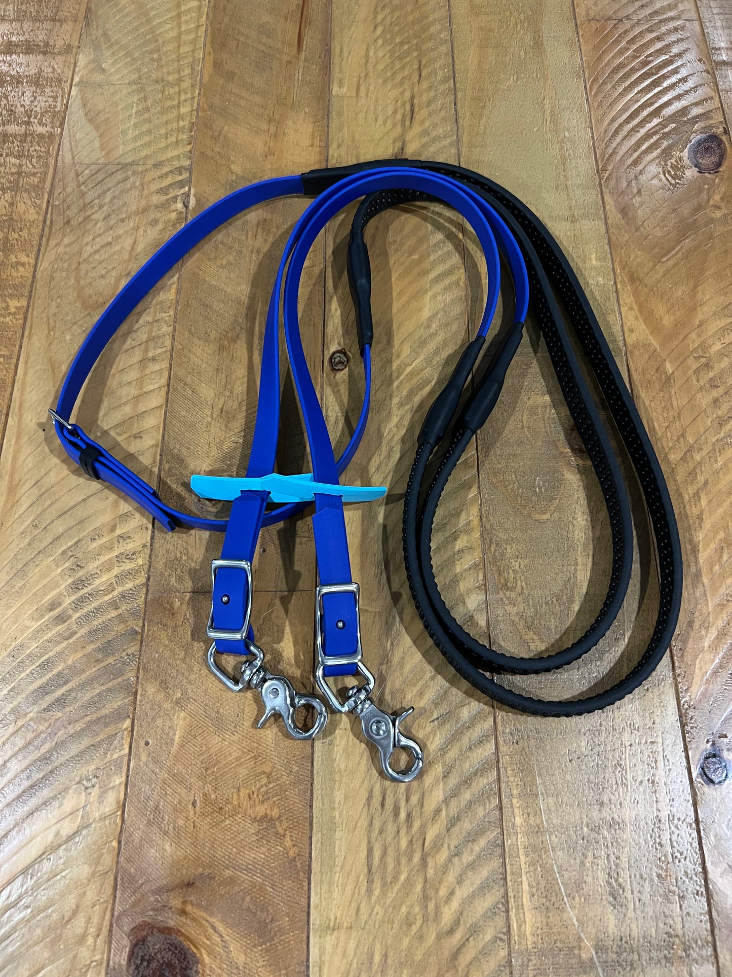 On a wooden surface, two dog leashes are displayed—one in blue and the other in black. Both leashes come equipped with metal clips for easy attachment to collars or harnesses. The blue leash, echoing the style of custom made bridles, features a turquoise tag and adds an elegant flair to your pet's outing. This product is known as "Soft Grip Reins - Royal Blue" by LS Equestrian.