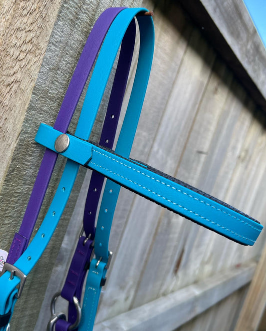 A close-up of the LS Equestrian's LS Deux Bridle - Lagune & purple, crafted from BioThane, hanging on a wooden fence. This bridle features a vibrant blue headpiece with purple and teal straps, accented with silver hardware. Ideal for endurance riding, its striking design contrasts beautifully with the clear sky above.