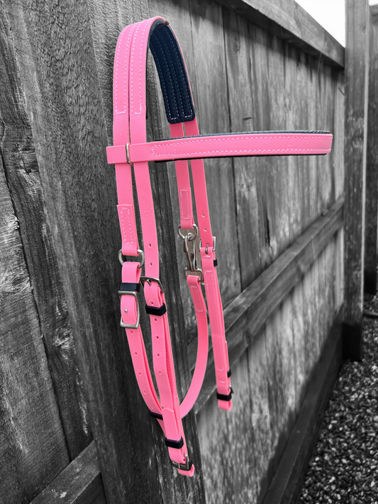 A striking Padded Race Bridle in Pastel Pink from LS Equestrian, product code PK522, hangs on a wooden fence. One adjustable strap is crafted in a contrasting darker hue, possibly blue. The grayscale background emphasizes the bridle's bright color and durable BioThane construction, while small gravel stones cover the ground, completing this vivid scene.