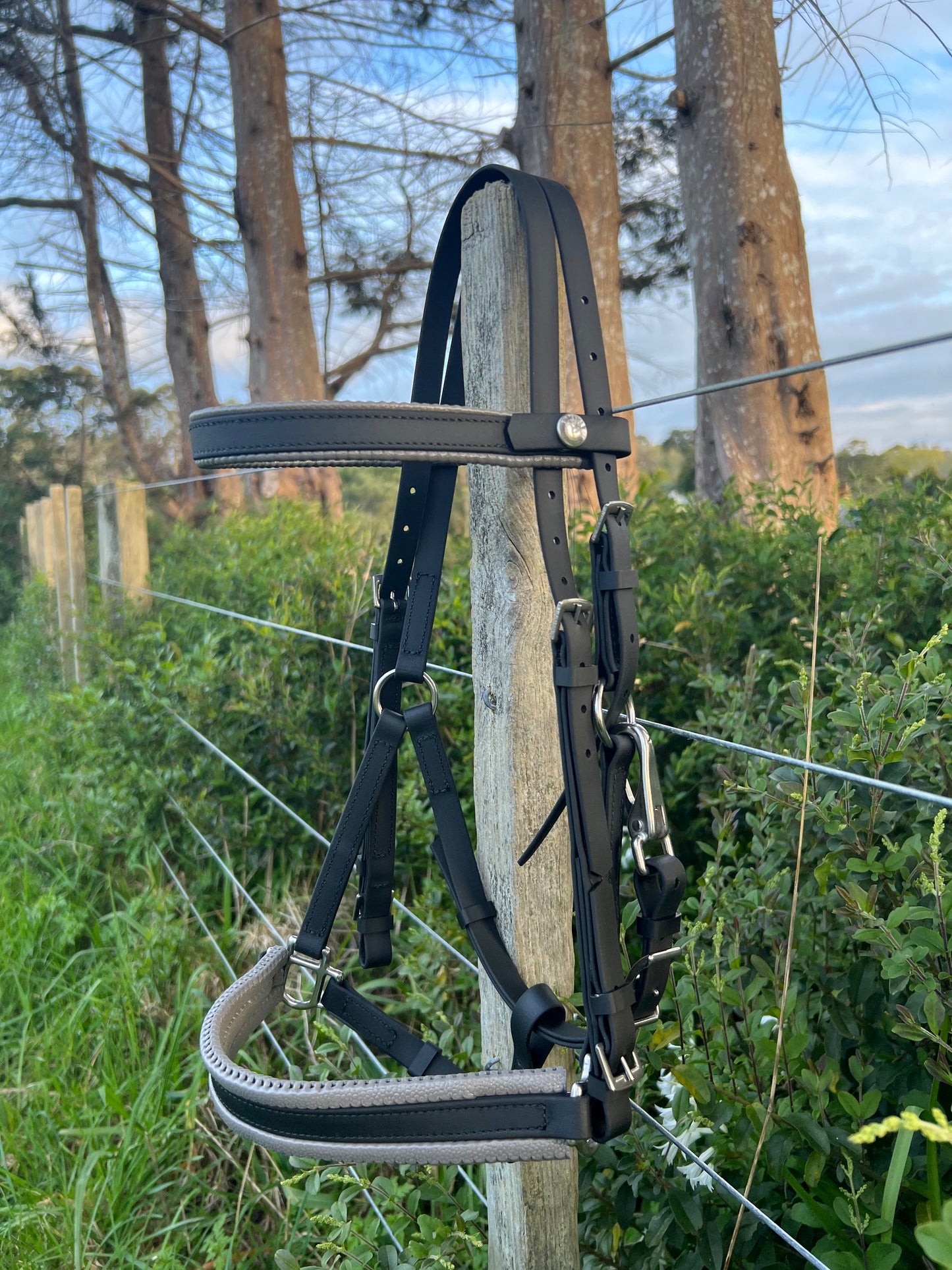 An LS Deux Bridle - Padded Black & Grey from LS Equestrian hangs on a wooden post next to a wire fence, with green bushes and tall trees in the background under a partly cloudy sky—ideal for endurance riding enthusiasts.