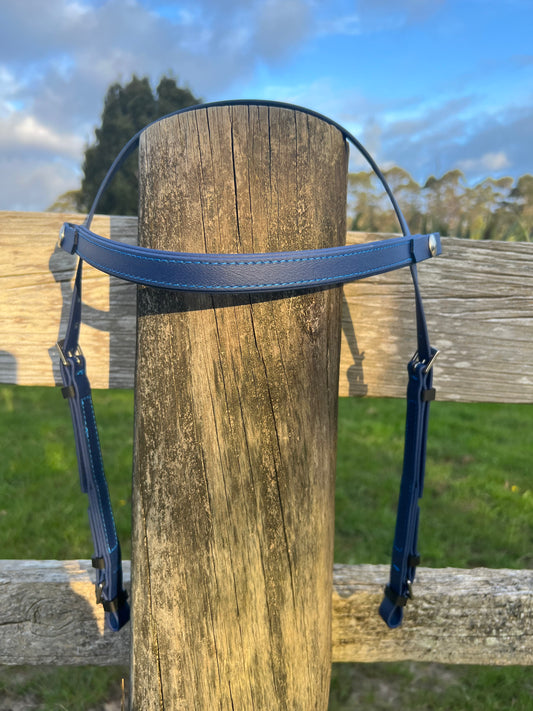 A navy blue LS Deux Headstall from LS Equestrian draped over a wooden fence post, with a backdrop of green grass and trees under a partly cloudy blue sky.