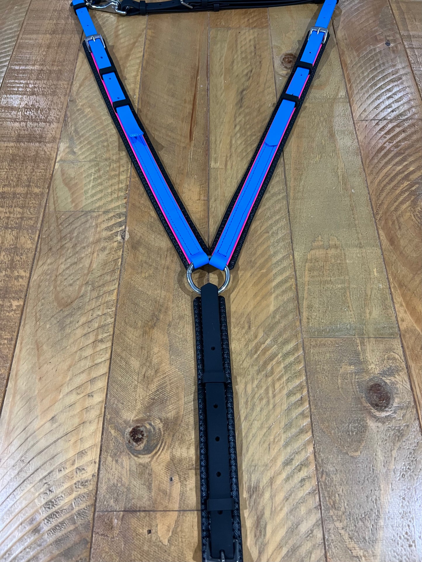 A striking, custom-crafted V-shaped harness from LS Equestrian in New Zealand is displayed flat on a wooden surface, showcasing cyan and hot pink straps with black borders. It's made with LS BETA® BioThane® and linked by an elegant metal ring.
