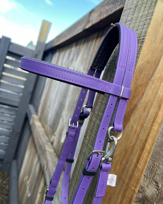 A violet purple Padded Race Bridle by LS Equestrian with sleek silver hardware contrasts against a wooden and metal fence, the sky peeking through in the top left corner, adding serenity to the scene.