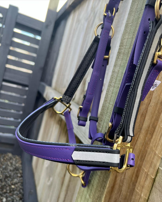 A Reflective BioThane®️ Halter - Padded design your own by LS Equestrian in purple and black, equipped with gold hardware, hangs on a wooden fence. The light brown fence is constructed from vertical panels, and a dark gate can be seen in the backdrop. Created with night riders in mind, the halter incorporates reflective strips for improved visibility and safety.