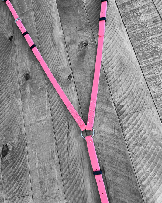 A Breastplate Zero Padding in coral pink leather, designed by LS Equestrian and featuring black buckles, is displayed on a rustic wooden surface, highlighting its unique style.