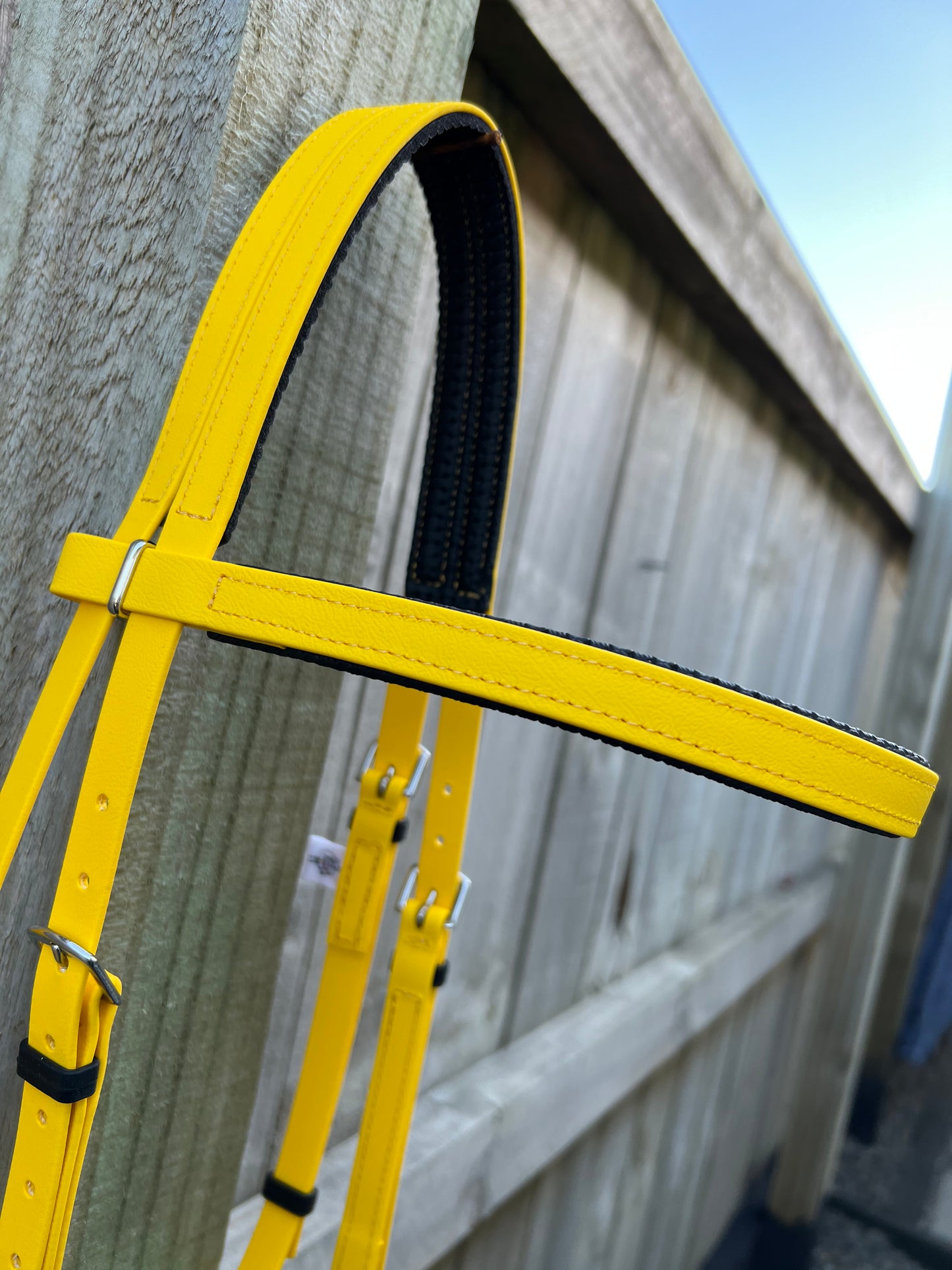 The LS Equestrian Race Bridle - Yellow, featuring a striking black lining, hangs elegantly on a wooden fence. Made from durable BioThane, this custom bridle perfectly complements the blue sky and blurred fence background.
