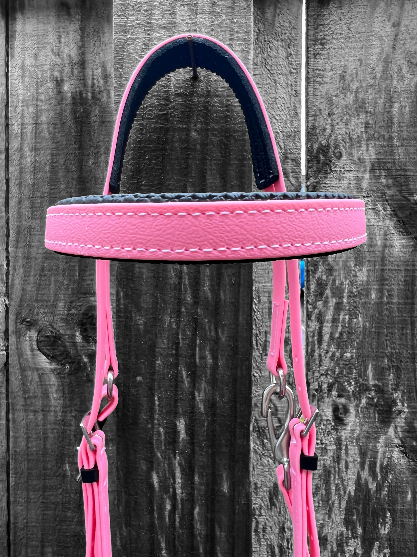 A Padded Race Bridle in Pastel Pink (PK522) by LS Equestrian, showcasing custom pink stitching and metallic hardware, hangs against a weathered wooden fence.