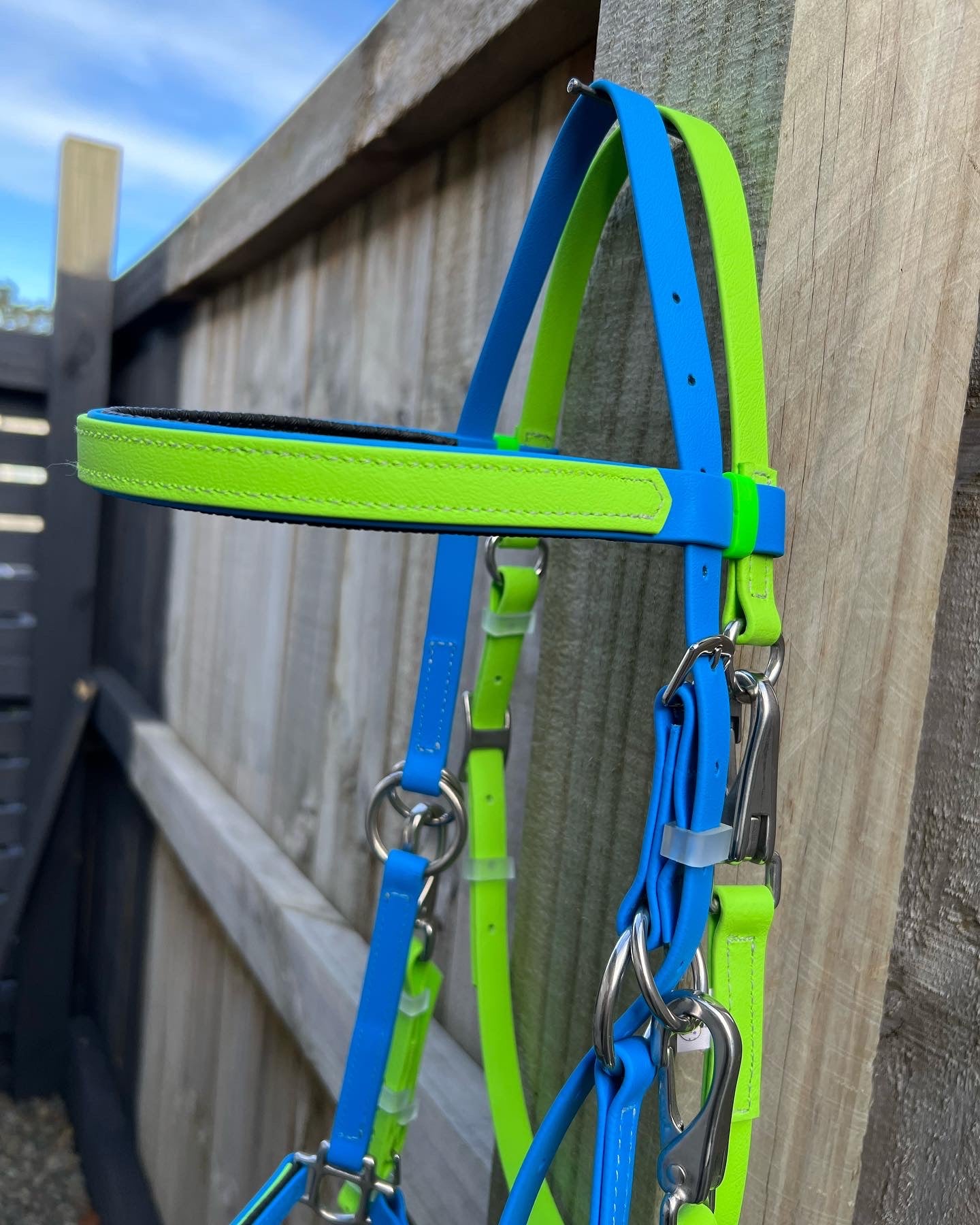 An LS Equestrian LS Enduro Bridle in apple and cyan hangs on a wooden fence, showcasing its custom design. The silver hardware provides a striking contrast against the colorful straps. Made in New Zealand, this piece sits under a clear blue sky.