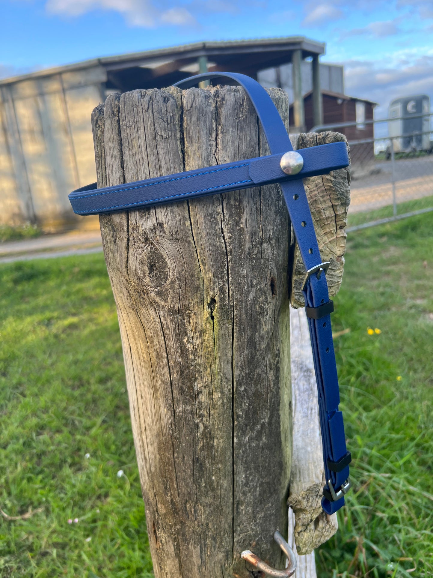 A navy blue LS Deux Headstall by LS Equestrian hangs on a wooden fence post, reminiscent of the craftsmanship seen in their Enduro range. In the background, a grassy area and a building sit under a partly cloudy sky.
