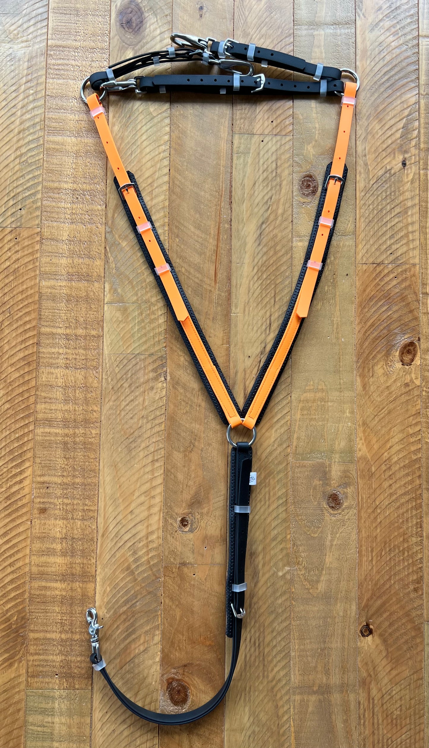 A Padded Breastplate in pastel orange and black from LS Equestrian rests on a wooden floor. Equipped with multiple metal buckles and rings for attachment, this design combines classic style with contemporary durability.