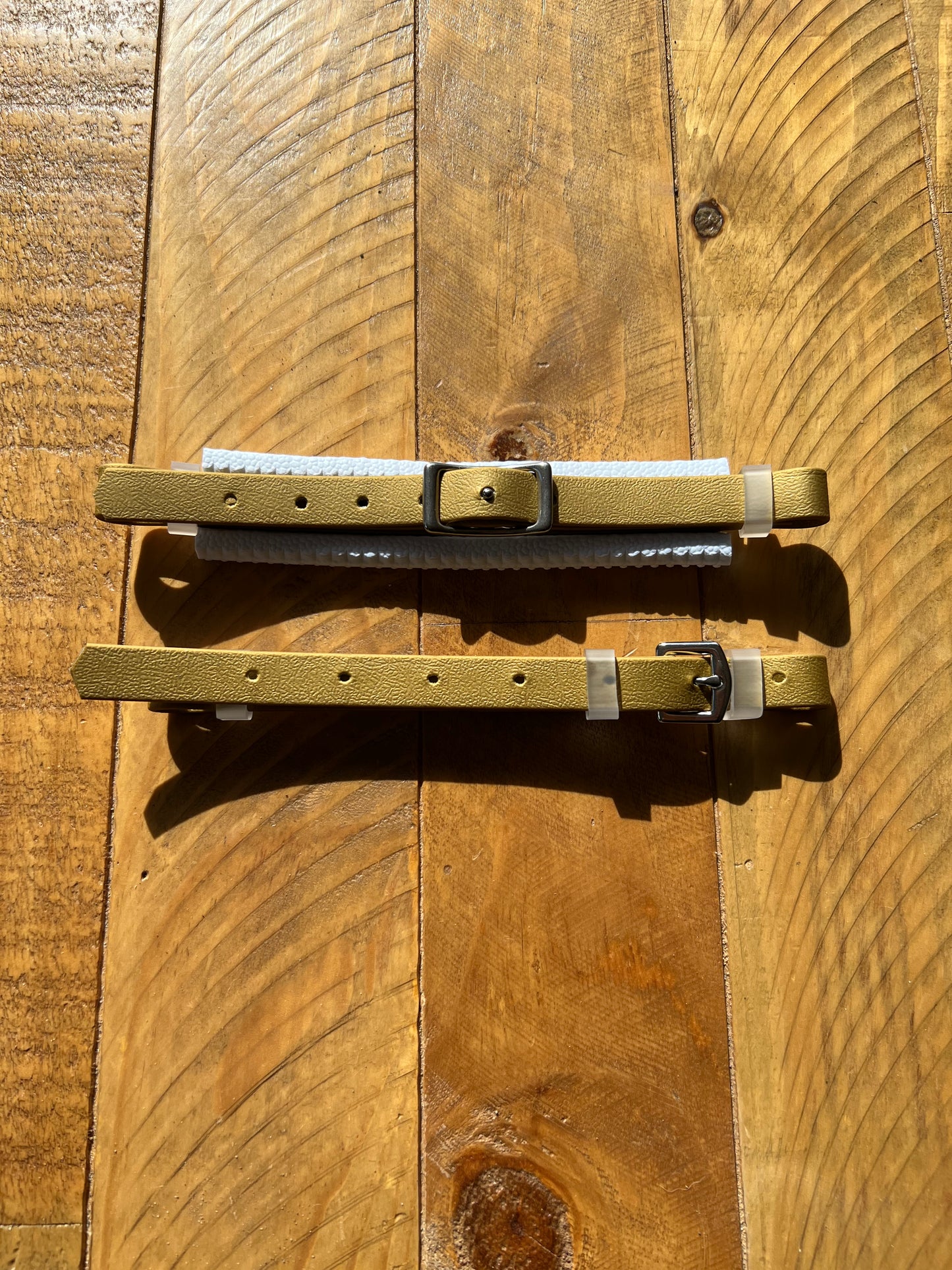 Two beige leather straps with black buckles are laid out on a wooden surface. Sunlight casts shadows, highlighting the texture of the wood and leather, reminiscent of fine craftsmanship synonymous with LS Equestrian's Hackamore Replacement Padded Nose Set.