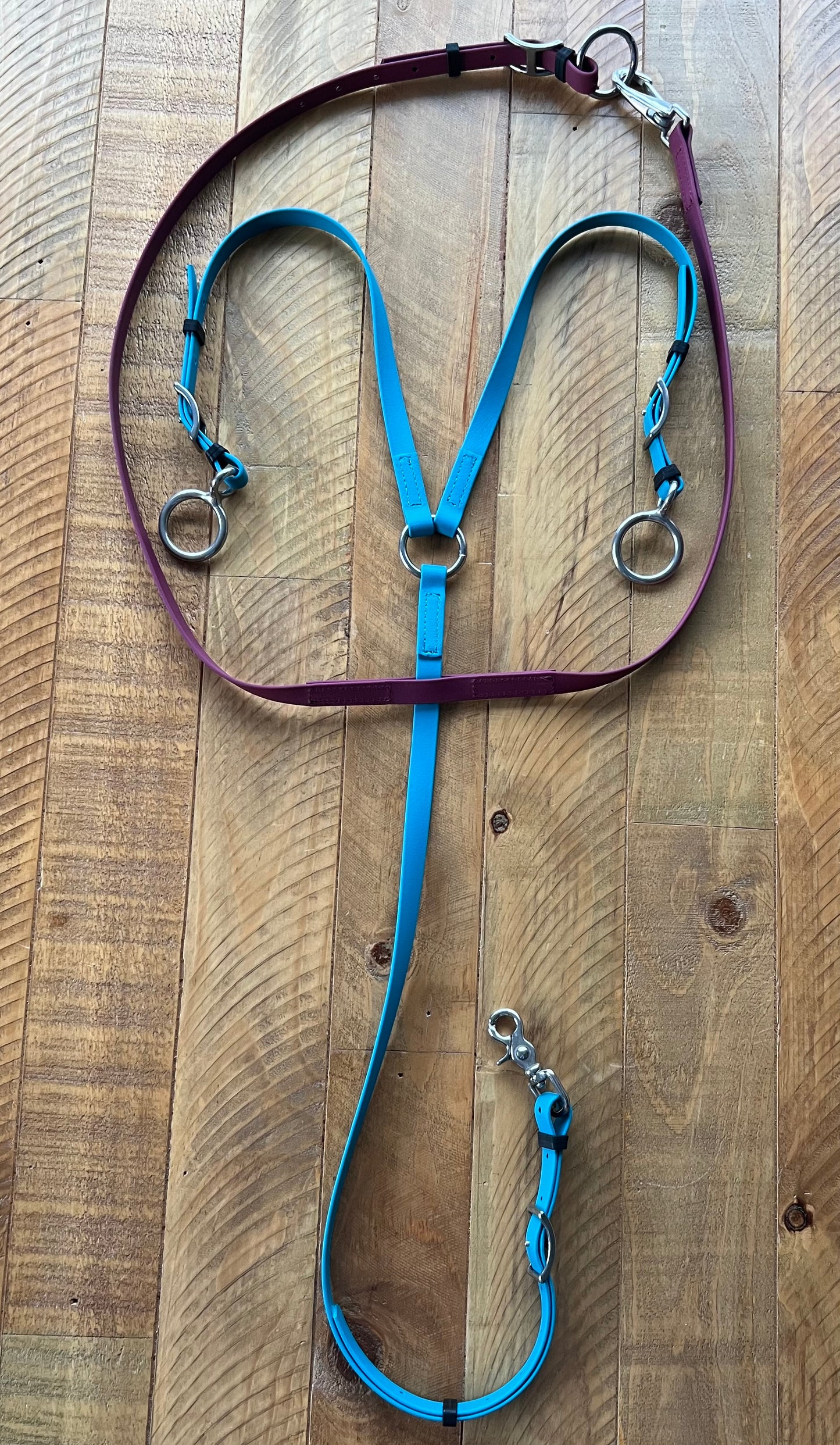 A vibrant Running Martingale from LS Equestrian, designed with durable Beta BioThane in a striking mix of purple and blue, enhanced with stainless steel rings and clips, is displayed on a wooden surface. The adjustable blue reins feature convenient clasps, making it ideal for any equestrian setup.