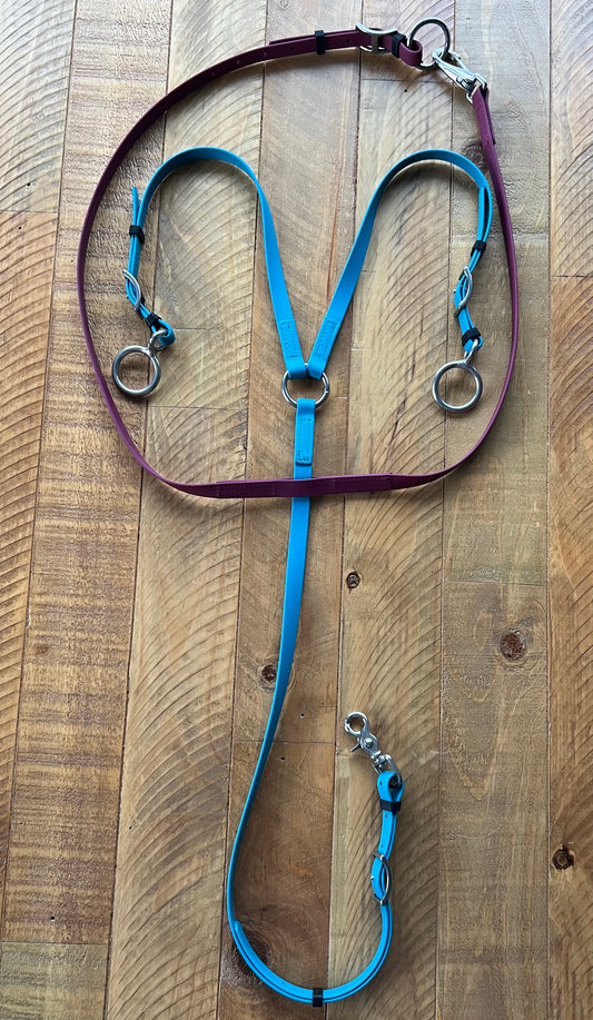 A vibrant Running Martingale from LS Equestrian, designed with durable Beta BioThane in a striking mix of purple and blue, enhanced with stainless steel rings and clips, is displayed on a wooden surface. The adjustable blue reins feature convenient clasps, making it ideal for any equestrian setup.