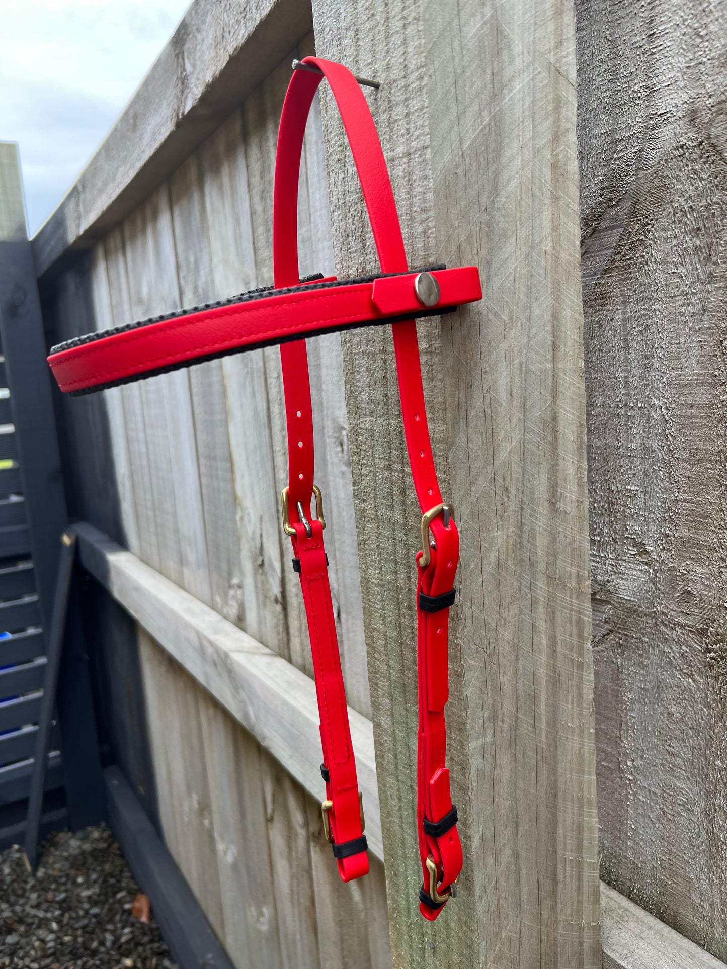 An LS Deux Bridle - Red from LS Equestrian, featuring advanced BioThane technology, hangs on a wooden fence. Its silver buckles shine against the smooth finish and textured strap, making it perfect for endurance riding. The sturdy vertical planks of the fence stand proudly outdoors.