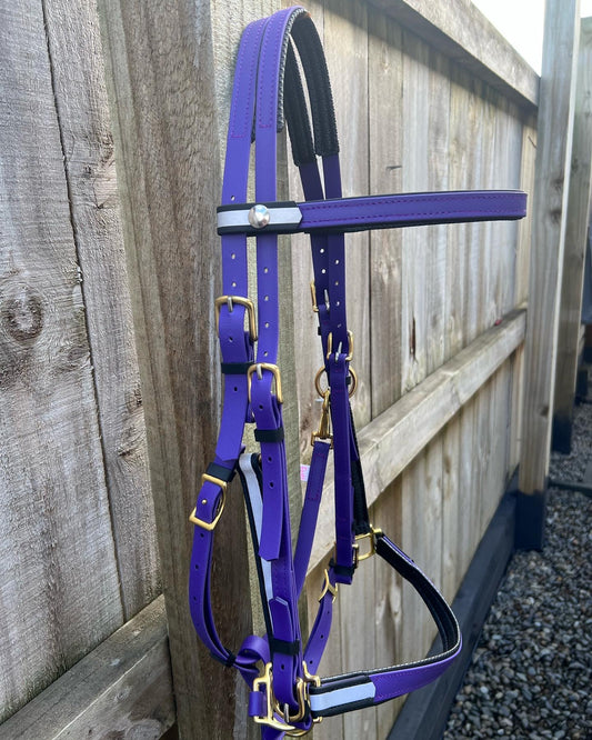 An LS Equestrian Reflective Ls Deux 2 Part Endurance Bridle, padded and custom-designed with durable BioThane in purple with gold buckles and black accents, hangs on a wooden fence against a gravel pathway.