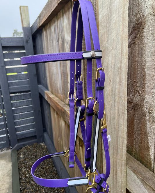 A vibrant purple custom LS Equestrian Reflective Ls Deux Bridle, complete with brass hardware, hangs on a wooden fence. This unique BioThane bridle features adjustable straps and silver buckles, perfectly complementing the wooden and metal fencing backdrop.