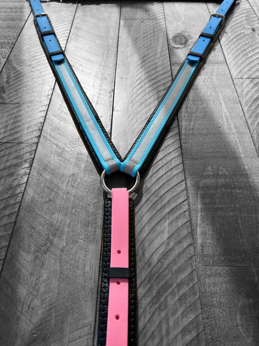 A close-up of the LS Equestrian Reflective Breastplate in light blue, cyan blue, and pastel pink laid out on a textured wooden surface. The harness features straps meeting at a metal ring in a Y shape and is BioThane® padded with BETA® finish. The grayscale background accentuates the vibrant colors of this meticulously crafted piece.