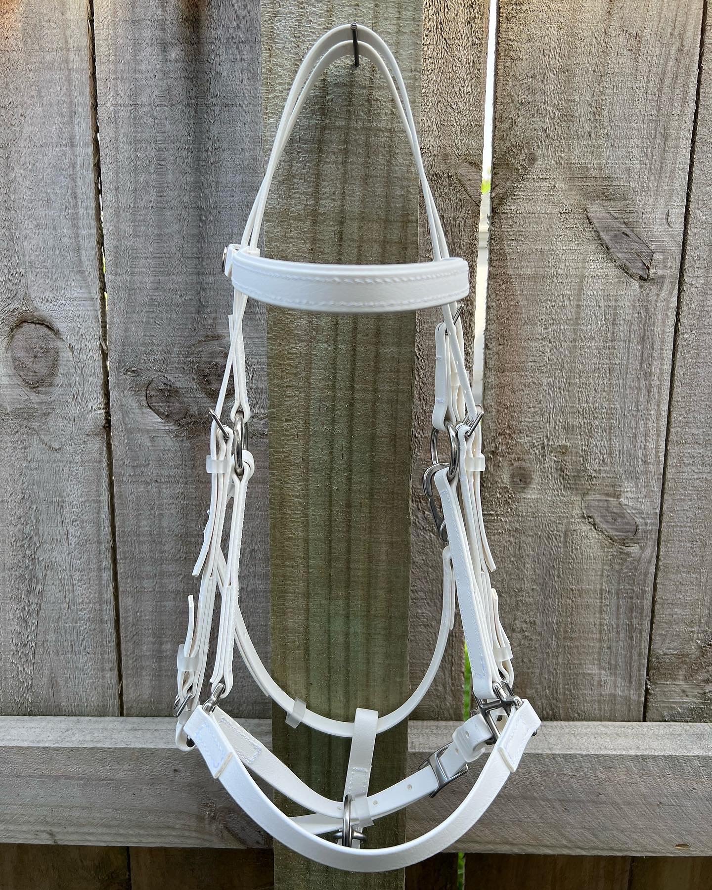 A white 2 Part Bridle from LS Equestrian is hanging on a wooden fence. The rustic wood, showcasing its visible grain and knots, beautifully complements the bridle's sleek design. Perfect for training, the straps and buckles are neatly displayed, making it ideal for endurance riders seeking both durability and style.