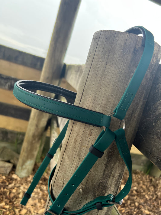 An LS Equestrian LS Barcoo Bridle - Design your own in green hangs on a wooden fence post, highlighting its position within the premium LS Endurance range. The background shows part of the fence and scattered woodchips on the ground, contributing a rustic charm to the scene.