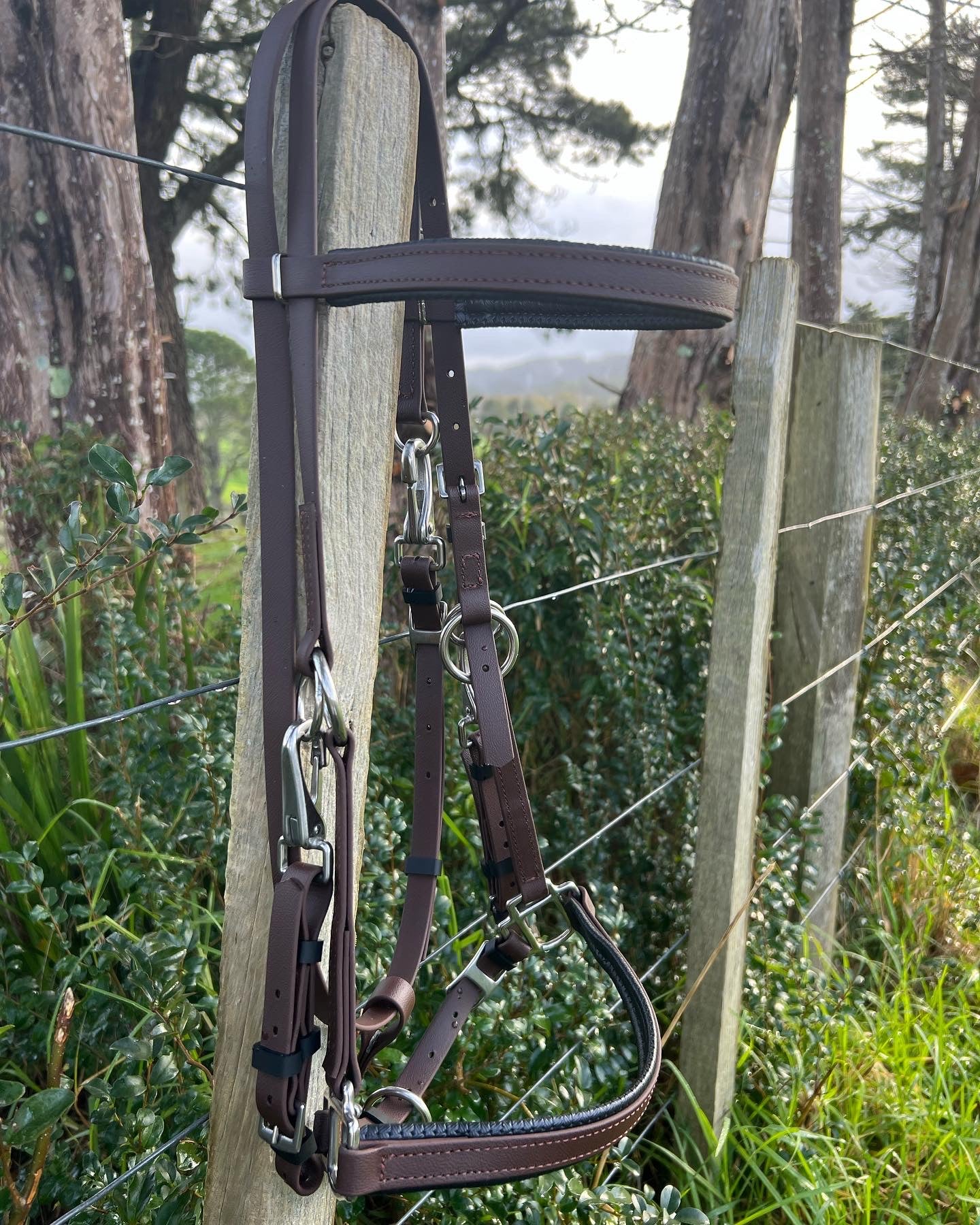 An LS Equestrian LS Enduro Bridle in dark brown, tailored specifically for endurance riders, is displayed hanging on a wooden fence post. The background showcases rich green foliage and several towering trees, enhancing the rustic, natural atmosphere.