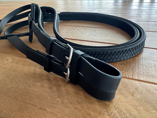 Close-up of custom Reins Pimple Grip belts by LS Equestrian, featuring black leather with silver buckles on a wooden surface. One has a textured pattern, the other is smooth. The image showcases craftsmanship and quality of these bespoke accessories.