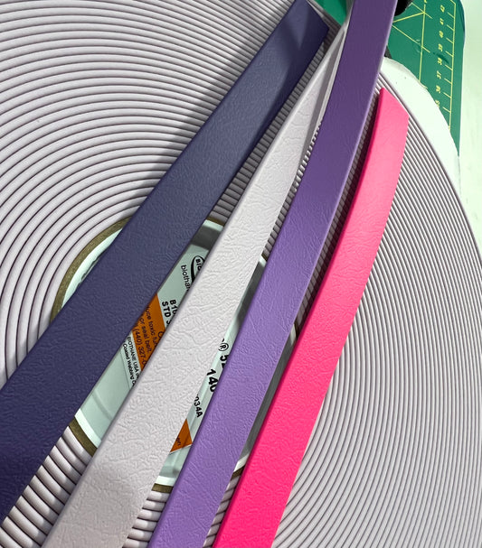 A close-up of the Beta®️ BioThane®️ Colour Sample Pack by LS Equestrian showcases a vibrant combination: five rolls of textured, colorful faux leather strips in shades of purple, lavender, white, pink, and magenta. They rest over coiled white piping on a cutting mat, enticing you to experience them firsthand.
