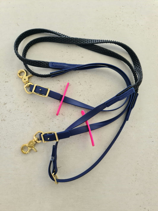 On a gray surface lie leather dog leashes in black and blue with gold hardware, evoking the style of custom-made LS Equestrian Soft Grip Reins in navy. Beneath the leashes rests a bright pink plastic stick.