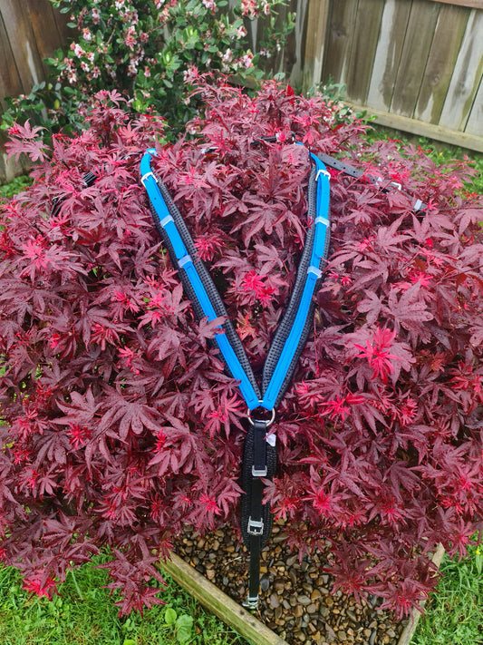 A Padded Breastplate in Cyan & Black by LS Equestrian, meticulously crafted with BioThane®, is elegantly draped over a small bush with deep red leaves, set in a wooden planter surrounded by grass. A wooden fence and more greenery provide the backdrop for this stylish scene.