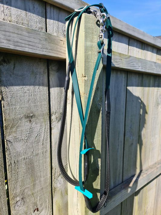 Two Soft Grip Reins in hunter green, crafted by LS Equestrian and equipped with sturdy metal clasps, hang on a weathered wooden fence in sunlight. Part of the LS Enduro range, these custom-made reins convey a sense of a sunny outdoor area.
