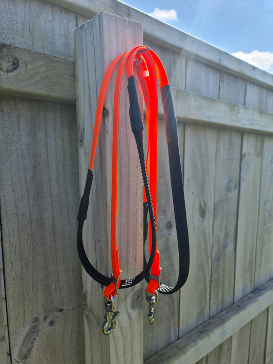 Two vibrant fluro orange soft grip reins, from the LS Equestrian brand, with metal clips hang on a wooden fence post. The custom-designed reins stand out vividly against the muted wood, with a clear sky and a few clouds in the background.