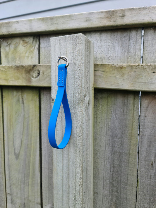 A blue Girth Loop from LS Equestrian, featuring a metal ring and girth loops, hangs from the top of a weathered wooden fence post with a natural finish.