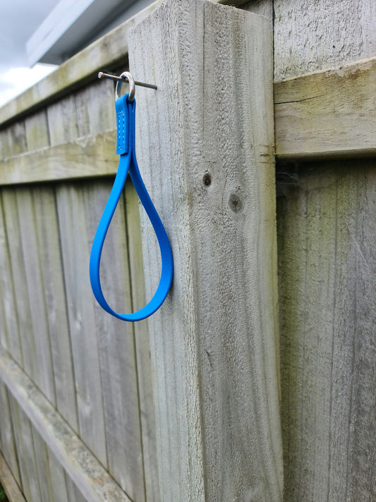 A Girth Loop by LS Equestrian, made of blue rubber, hangs from a nail on a weathered wooden fence, with an overcast sky in the background.