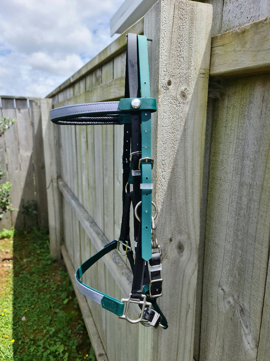 A leather LS Deux Bridle in hunter green, black, and grey from LS Equestrian hangs on a wooden fence outdoors. The sky is partly cloudy, with green grass and a small bush visible below the fence, capturing the essence of endurance riding scenery.