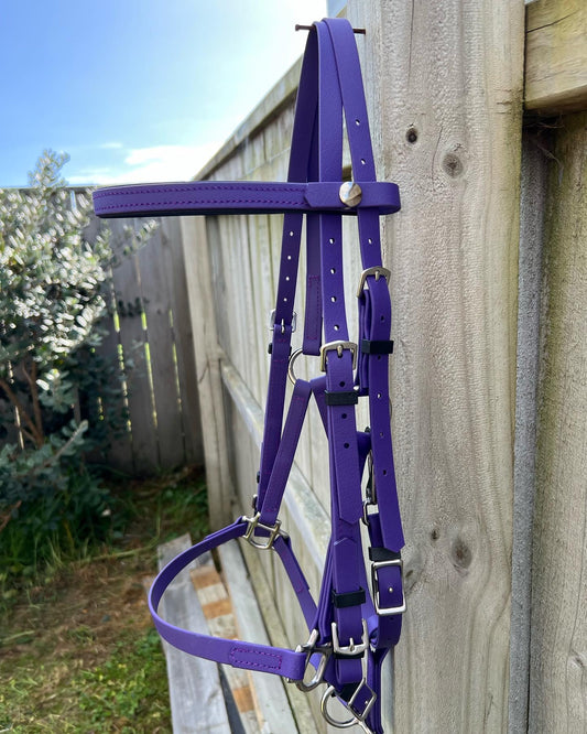 An LS Deux Bridle - Violet Purple by LS Equestrian, crafted in New Zealand, is draped over a wooden fence outside, surrounded by vibrant greenery in the background.