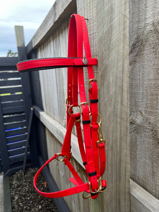 The LS Deux Bridle - Red by LS Equestrian, ideal for endurance riding, drapes over a wooden fence. It boasts multiple adjustable straps with silver buckles and brass-colored rings. In the background, more wooden fences with horizontal panels can be seen.