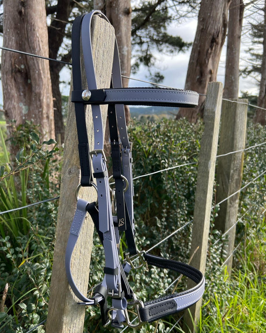 An LS Equestrian LS Deux Bridle - Padded Grey & Black is hanging on a wooden fence post. The background features a wire fence, green bushes, and several trees under a cloudy sky, ideal for some endurance riding.