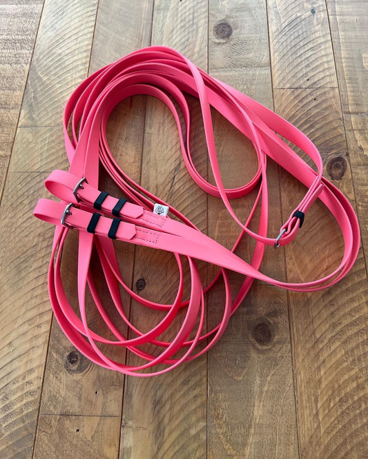 A pair of coiled coral driving reins made from BioThane® by LS Equestrian, featuring two buckles and black accents, lies on a wooden surface.