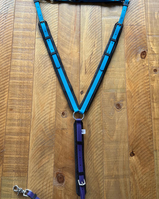The Padded Breastplate - Lagune blue & purple from LS Equestrian is displayed laid flat on a wooden surface. The BioThane design showcases a Y-shape with adjustable straps and a durable breastplate, which includes a metal ring at the center. Additionally, there's a visible leash attachment at the bottom.