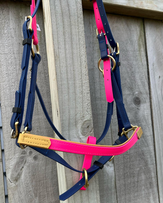 The LS Deux Bridle by LS Equestrian, handmade in New Zealand, showcases vibrant hot pink, navy, and gold straps with brass hardware. It hangs elegantly on a wooden fence.