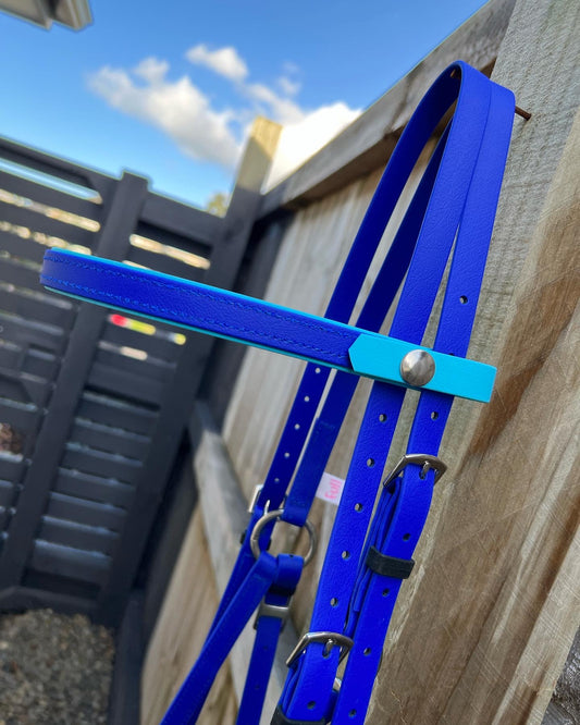 A close-up of an LS Equestrian LS Deux Bridle in striking royal blue and light blue hues hangs on a wooden fence. The backdrop showcases vertical slats and a partly cloudy sky, capturing the essence of endurance riding from the LS Enduro collection.