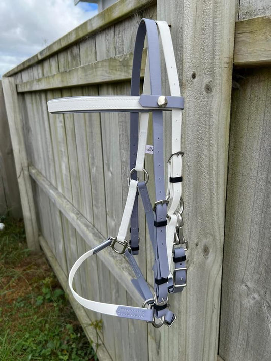 An LS Deux Bridle in White & Grey from LS Equestrian, featuring metallic buckles and loops, is draped over a wooden fence, framed by a scene of lush green grass and an overcast sky.