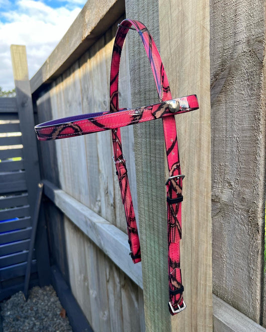 A vibrant pink and black patterned LS Equestrian Bridle Headstall - Camo hangs gracefully on a wooden fence under a partly cloudy sky.