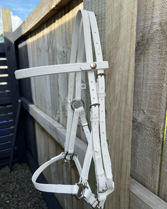 A white 2 Part Bridle from LS Equestrian, featuring shiny metal buckles, is hanging on a wooden fence outside. The scene is set against a clear sky with a glimpse of a garden area, making it perfect for endurance riders gearing up for their next adventure.