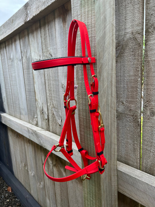 An LS Equestrian LS Deux Bridle in vibrant red, ideal for endurance riding, with elegant gold hardware, is draped over a weathered wooden fence. The gravelly ground beneath adds to the rustic setting. This bridle includes multiple adjustable straps and buckles to ensure comfort during long rides.