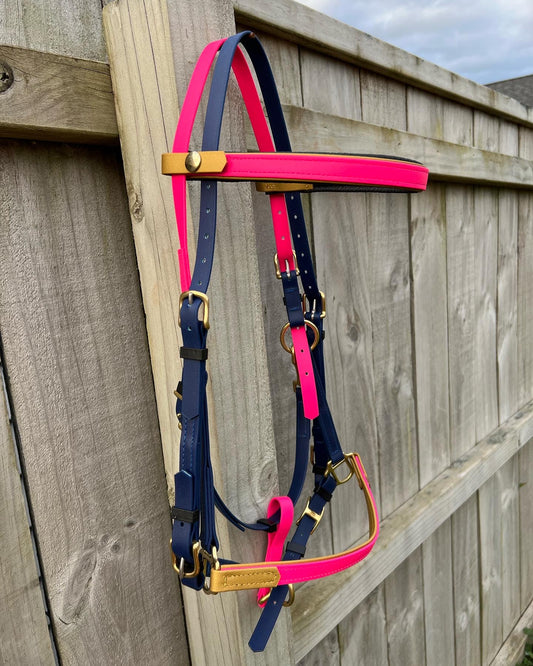 A LS Deux Bridle in Hot Pink, Navy & Gold from LS Equestrian, made with Beta BioThane, adorns a wooden fence. The bridle's gold buckles and accents create a striking contrast against the natural wood. Above, an overcast sky looms.