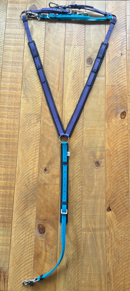 The "Padded Breastplate - Purple & Lagune Blue" by LS Equestrian rests on a wooden surface, showcasing its adjustable BioThane® straps in striking blue and purple with metal rings and buckles, crafted into a Y-shape.
